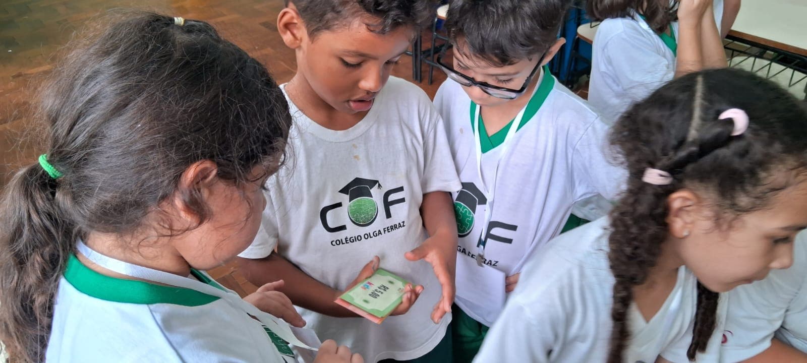 Grupo de alunos reunidos e inclinados sobre uma cédula de cinco reais do material, que um deles segura aberta na mão.
