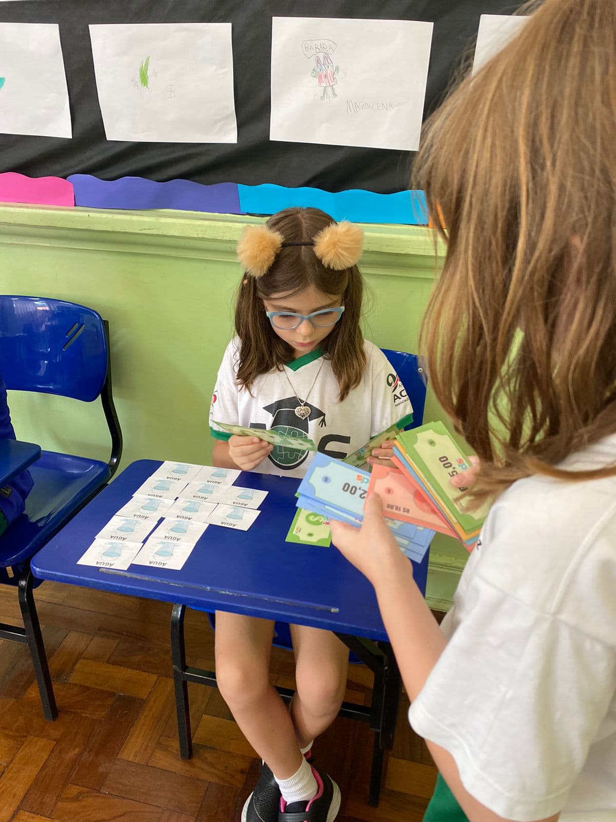Aluna sentada na carteira examina uma cédula do material, com cartões ilustrados de água à sua frente e, ao fundo, um painel com desenhos feitos pelos alunos.