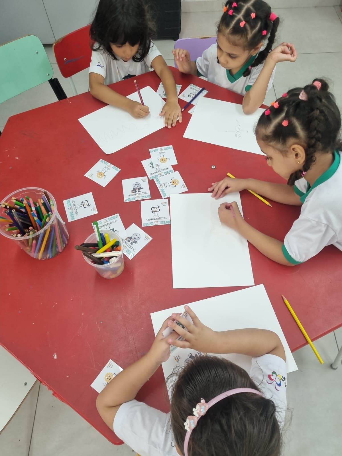 Quatro crianças desenham em folhas brancas em volta de uma mesa vermelha, com potes de lápis e cartas dos personagens DINI, Senhor Guardião, Contazinha e Trocazinha espalhadas entre elas.