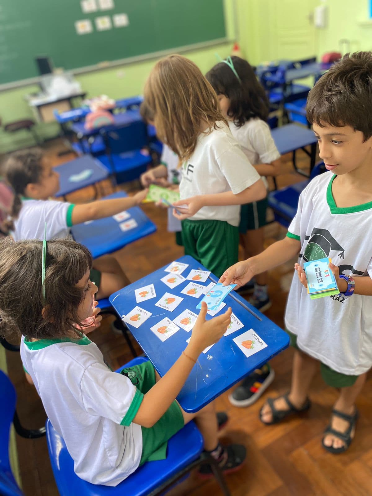 Vários alunos circulam entre as carteiras da sala trocando cédulas do material por cartões ilustrados de cenoura.