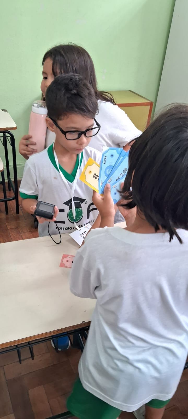 Aluno de óculos segura uma maquininha de cartão de brinquedo em uma mão e cédulas de dois reais do material na outra, atendendo um colega do outro lado da mesa.