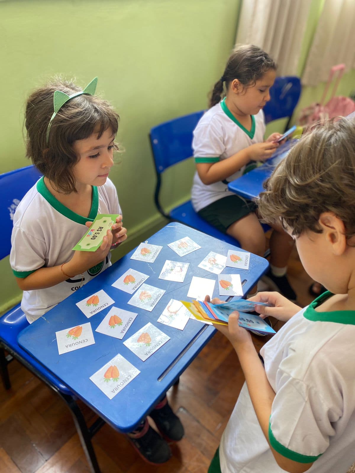 Alunos em volta de carteiras com cartões ilustrados de cenoura espalhados sobre a mesa, segurando cédulas coloridas do material da metodologia.