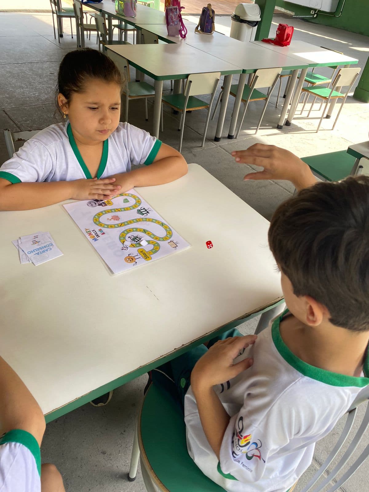 Dois alunos frente a frente em uma mesa, com o tabuleiro de trilha Money Skills Kids impresso entre eles, um dado vermelho e um monte de cartas-conselho.