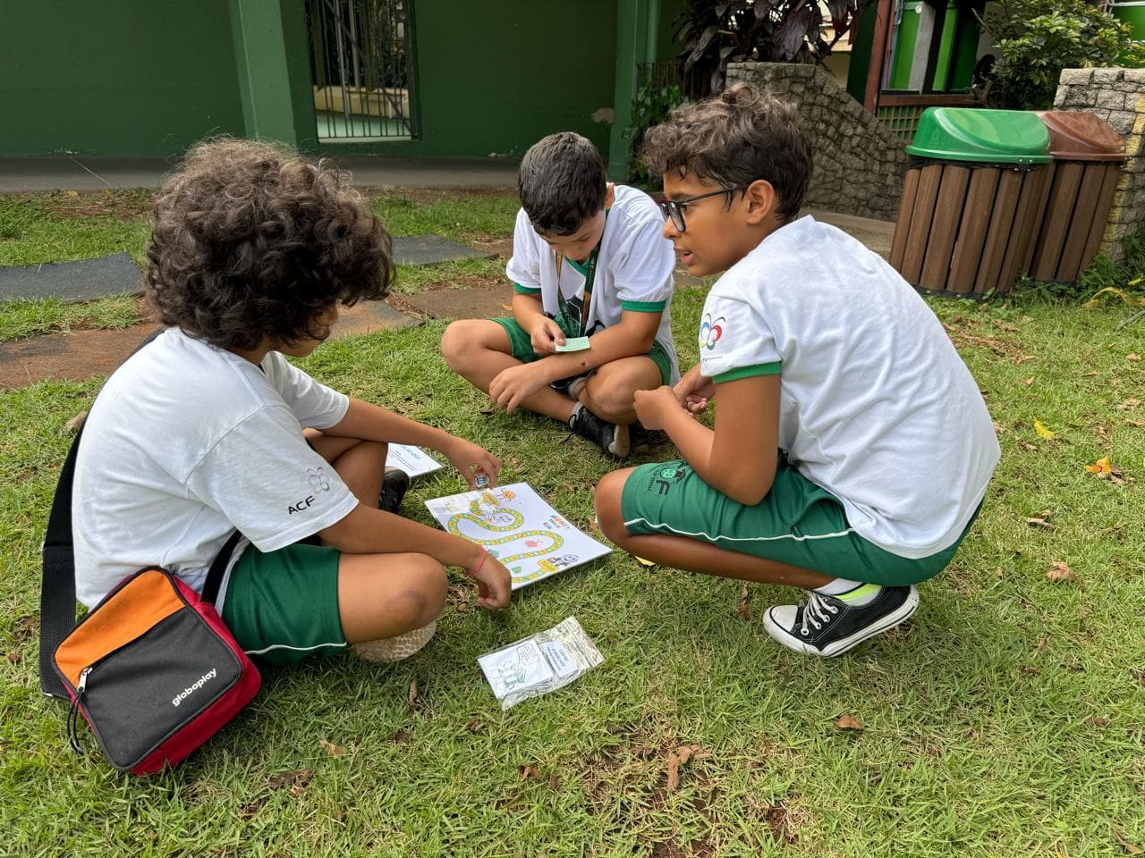 Três alunos de uniforme sentados na grama do pátio, em roda, em volta de um tabuleiro de trilha impresso, com cartas e cédulas do material espalhadas.